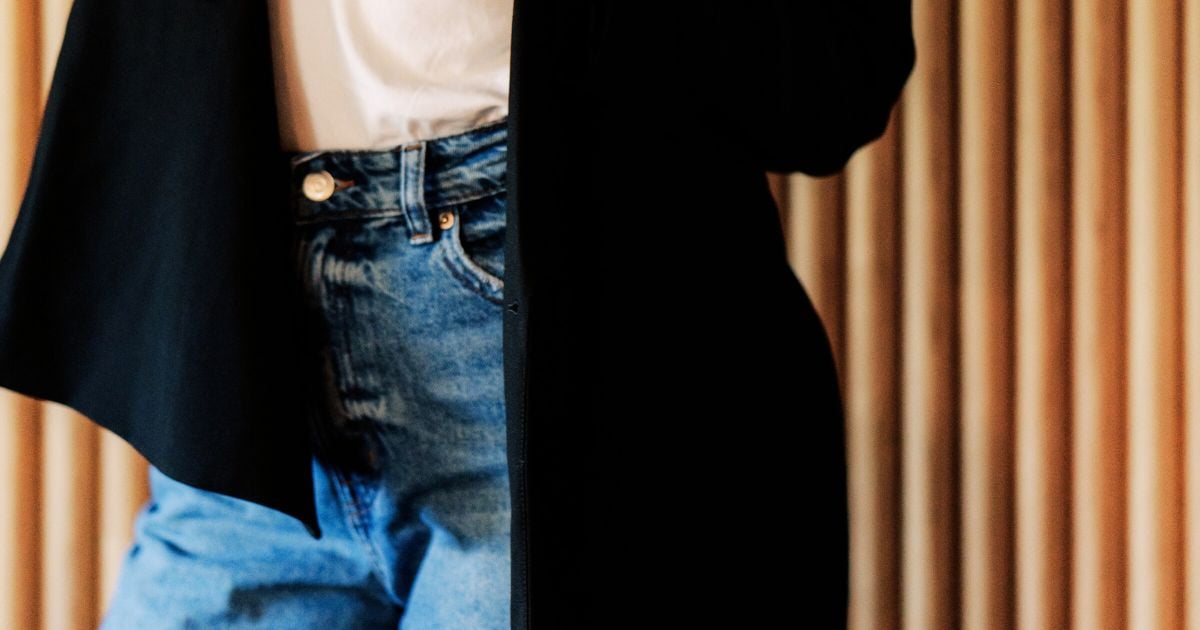 Lady leaning against a wood panelled wall wearing blue jeans, a white t shirt and a black blazer