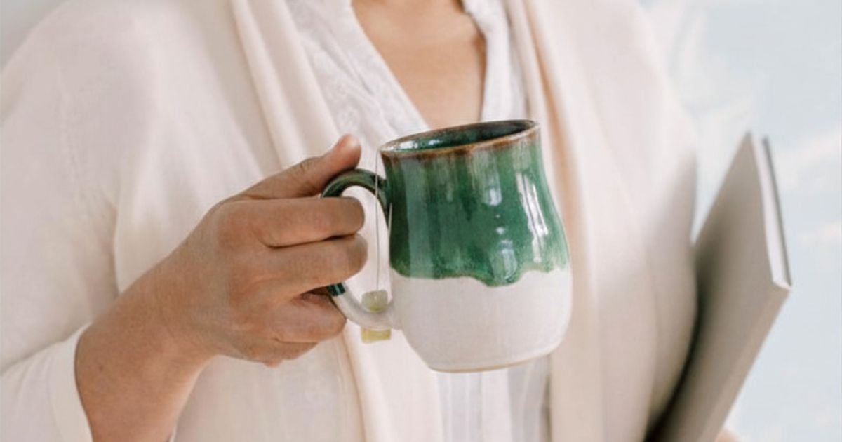 Lady holding a green and cream mug of tea in one hand and a planner in the other hand, smiling